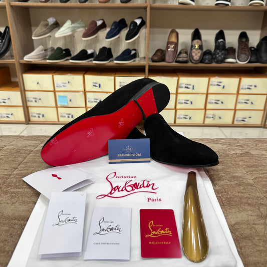 Christian Louboutin black suede dress shoes displayed with a red sole, branded box, care instructions, and a shoe shine in a retail showroom setting.