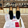 A pair of Christian Louboutin black suede slip-on shoes displayed on a white branded cloth with shoe boxes, a gold shoe tree, and brand cards in front of a shoe rack in a retail store.