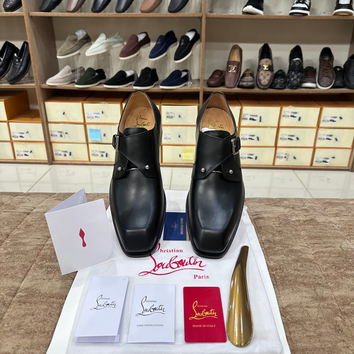 A pair of Christian Louboutin black leather double-strap loafers displayed on a white cloth with brand accessories, including a shoe box, cards, and a gold shoe shine, in front of a shoe retail display.