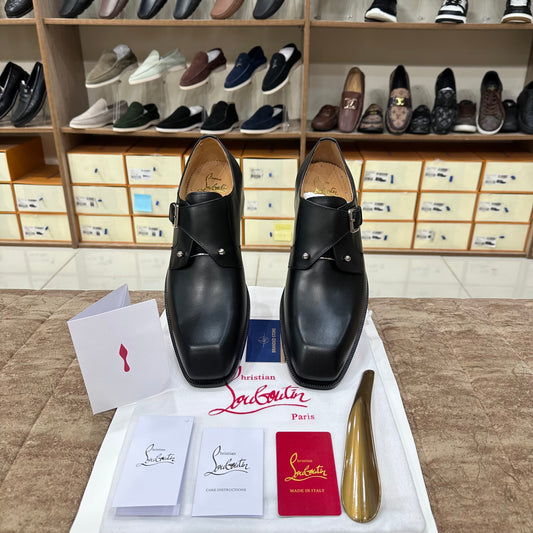 A pair of Christian Louboutin black leather double-strap loafers displayed on a white cloth with brand accessories, including a shoe box, cards, and a gold shoe shine, in front of a shoe retail display.